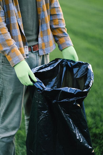 A person wearing a casual plaid shirt in shades of yellow, blue, and gray, paired with green gloves, is holding open a large black plastic rubbish bag outdoors on a grassy area. The bag appears to be partially filled and is ready for rubbish disposal or collection. The environment suggests a garden or park setting, with a blurred background of green grass. The individual's midsection, arms, and hands are visible, with the person standing upright and slightly leaning forward as they hold the bag open. This scene reflects aspects of waste management or rubbish collection, typical of activities undertaken by private waste handling services such as those offered by companies like rubbishcollectionwesthampstead.co.uk, aimed at efficient on-site clearance and rubbish disposal.