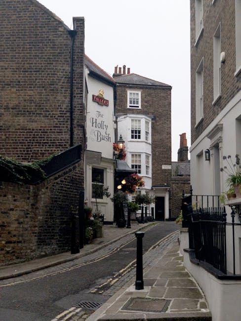 A narrow residential street in what appears to be an older urban area, showing multiple interconnected buildings constructed from brick and rendered materials, with varying architectural styles. In the foreground, a small section of cobblestone pavement and a metal bollard are visible, guiding pedestrians along the footpath. On the left, a brick wall partially covered with greenery curves around a corner, leading to a white building with a sign reading 'The Holly Bush' and some hanging flower baskets and potted plants outside its entrance, indicating a possible pub or inn. To the right, a white-painted building with large, multi-pane windows and decorative railings on a small balcony is visible, as well as a wall-mounted outdoor light. The background reveals additional brick buildings with chimneys and slate roofs, under a cloudy sky, suggesting a typical historic district scene that might be serviced by private waste collection or rubbish removal providers like rubbishcollectionwesthampstead.co.uk to manage waste disposal outside usual municipal services.