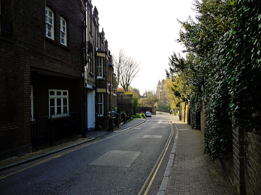 A quiet residential street lined with dark brick terraced houses on the left, each with small front gardens enclosed by black metal fences. The pavement on the right is paved with concrete tiles and bordered by a high, dense hedge of evergreen foliage, casting partial shade. The asphalt road slopes gently downward and features double yellow lines along the edge, indicating parking restrictions. In the distance, a white car is parked alongside more trees, some with bare branches suggesting early spring or late autumn. The scene is lit by natural daylight, with a bright, slightly overcast sky overhead, creating a neutral, unobtrusive atmosphere. The surroundings are clean and well-maintained, with no visible rubbish or debris, illustrating typical private residential waste management in an urban environment.