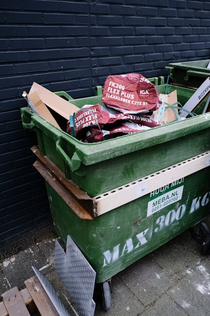 A green wheelie bin filled with red and black bags of construction or building waste, placed on a paved surface beside a dark blue brick wall. The bags appear to contain materials such as insulation or packing foam, with visible text including 'FK200 FLEXY PLUS' and 'FLEXLEIBER CZTE S1'. The green bin is slightly overflowing, with some bags leaning against the waste container's interior. The scene suggests a private waste disposal or alternative rubbish collection method, possibly handled by a specialist waste removal service like rubbishcollectionwesthampstead.co.uk, and is situated next to other waste bins and a wooden pallet leaning against the wall nearby.