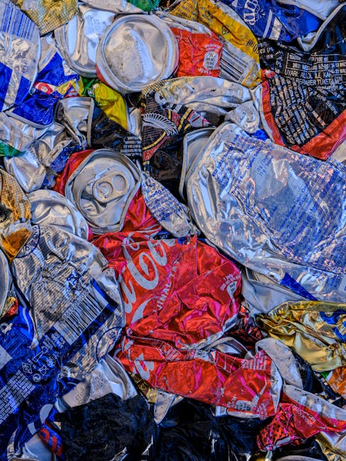 A close-up view of a large pile of crushed aluminum beverage cans, predominantly silver with various branding labels and some colored sections, showing a mix of crumpled and flattened textures. The cans are layered atop each other, with some partially visible labels from brands such as Coca-Cola, in red and white, along with other identifiable branding in black, blue, and yellow. The cans are resting on a surface that is not visible, suggesting they are discarded or prepared for collection. The lighting highlights the metallic sheen and creases in the cans, creating reflections and subtle shadows that emphasize their varied shapes and sizes. This scene exemplifies waste material typically associated with beverage containers awaiting recycling or rubbish removal, relevant to independent waste handling services or private disposal options, as seen in a waste management context.