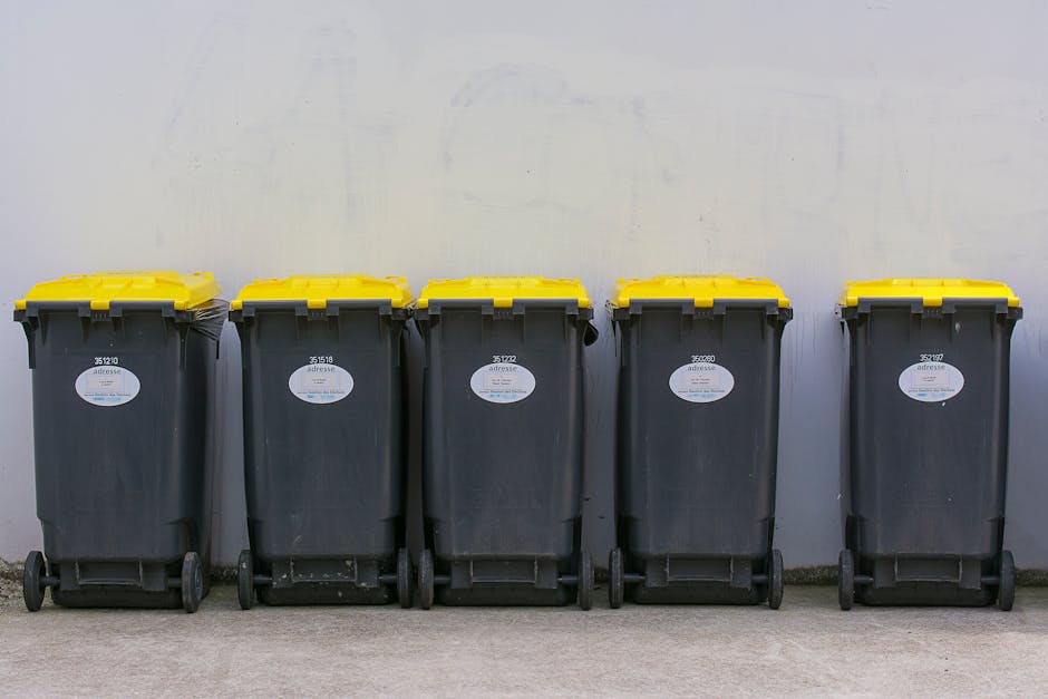 A row of five large wheeled rubbish bins is positioned against a plain, light grey wall, set on a concrete pavement. Each bin is made of durable black plastic with a textured surface, and all feature yellow lids with a smooth, glossy finish. The bins are arranged side by side, with the first four aligned closely and the fifth slightly offset to the right. Small white labels with barcodes and numbers are affixed to the front of each bin, indicating identification or collection details. The environment appears to be an outdoor storage area or service yard, with neutral lighting that highlights the clean, utilitarian design of the bins. This setup reflects an alternative waste management approach, such as private rubbish collection or on-site clearance, emphasizing responsible and organized rubbish removal. The scene is straightforward, functional, and typical of private waste handling services like those provided by companies specializing in rubbish removal for residential or commercial properties in NW6, West Hampstead.