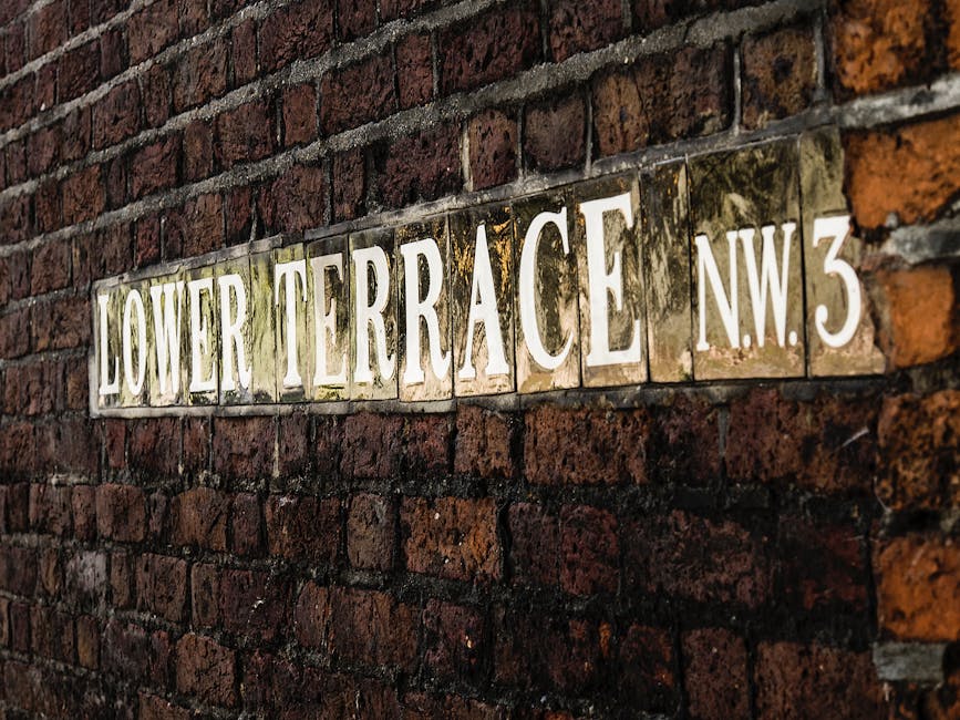 A close-up photograph of a rectangular street sign mounted on a weathered red brick wall. The sign displays the words 'LOWER TERRACE' and the additional identifier 'N.W.3' in large, white uppercase letters on a reflective, metallic background. The brick wall behind the sign features rough, uneven mortar joints and bricks with varying shades of dark red, brown, and black, some showing signs of weathering and moss growth around the edges of the sign. The photograph is taken at an angle, with the sign positioned horizontally across the frame, emphasizing the texture of the brick surface and the slightly aged appearance of the sign. The environment appears to be outdoor, with natural lighting highlighting the textures and color contrasts between the bricks and the sign, which could be part of an alleyway, residential street, or service area associated with private waste disposal or rubbish collection services aligned with independent rubbish removal.