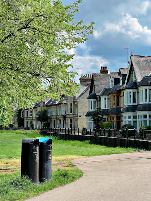 A paved pathway in a residential area lined with green grass and a black wrought iron fence in front of a row of traditional terraced houses with brick and stone facades. The houses feature bay windows, some with decorative wooden trims, and have pitched roofs with chimneys. To the left, a large deciduous tree with fresh green leaves extends its branches over the scene, creating partial shade. Two black public waste bins are positioned near the pathway on the grass, appearing slightly weathered with minor signs of use. The sky above is mostly blue with a few scattered white clouds, and there is natural daylight illuminating the scene. The image subtly showcases an environment where private waste management or rubbish collection services, such as those provided by companies like rubbishcollectionwesthampstead.co.uk, might be involved in maintaining cleanliness in residential neighbourhoods, especially in the context of alternative or independent waste disposal methods, without explicit promotional content.