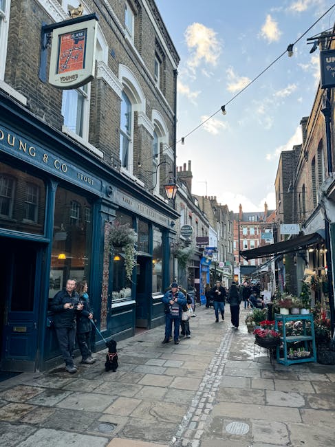 A narrow pedestrian street in an urban area featuring a mix of commercial shops and outdoor seating. On the left side, there is a building with a dark blue façade and large windows displaying interior furnishings, likely a café or pub, with a sign that reads 'Young & Co.' Above, a hanging illuminated sign advertises a Young's pub. Several pedestrians are walking along the uneven stone paving, some holding drinks or bags, and two individuals near the building are engaged in conversation. One person has a small black dog on a leash. To the right, there are small market stalls displaying potted plants and flowers, supported by a black canopy extending from a building. Overhead, a string of small outdoor lights runs across the street, and the sky above is partly cloudy with patches of sunlight. The scene suggests a lively, mixed-use area that may be targeted for private waste collection or rubbish removal services, fitting within an independent disposal or on-site clearance context through local providers like rubbishcollectionwesthampstead.co.uk.