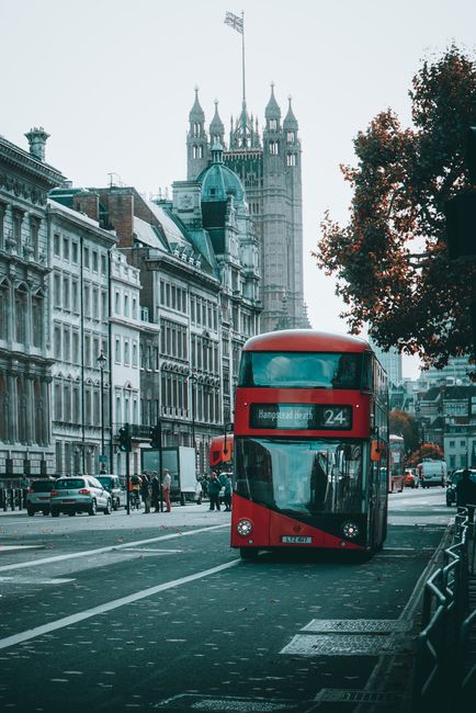 A red double-decker bus traveling along a city street with historic buildings lining the road and the Palace of Westminster with its iconic clock tower visible in the background. The bus displays route number 24 and is heading toward Hampstead Heath. Pedestrians are walking on the pavement, and several parked cars are visible alongside the road. The scene is captured during daylight with overcast weather, and trees with autumn foliage are seen on the right side of the image. This urban setting reflects active public transport and busy city life, which may be contrasted with private waste management or rubbish removal services that operate behind the scenes to maintain cleanliness in such environments. The image conveys a typical London street scene with historic architecture and a focus on city infrastructure, supporting the context of alternative waste handling methods such as independent collection or on-site clearance, supported by local or private providers like rubbishcollectionwesthampstead.co.uk, although the bus and street scenery are central to the visual narrative.